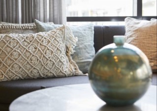 Detail shot of a bright lounge area. A modern vase is standing on a small, round table. The couch in the background is decorated with a selection of pillows.