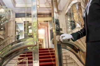 Eine weiß behandschuhte Hand öffnet eine große Glastür zu einem beeindruckenden Hoteleingang.